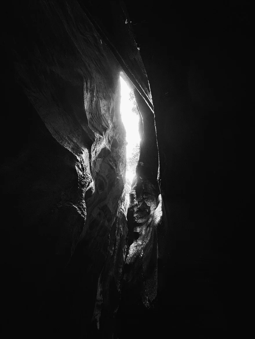 A narrow crevice of rock, almost entirely in shadow, with a single sliver of bright daylight cutting through the centre.