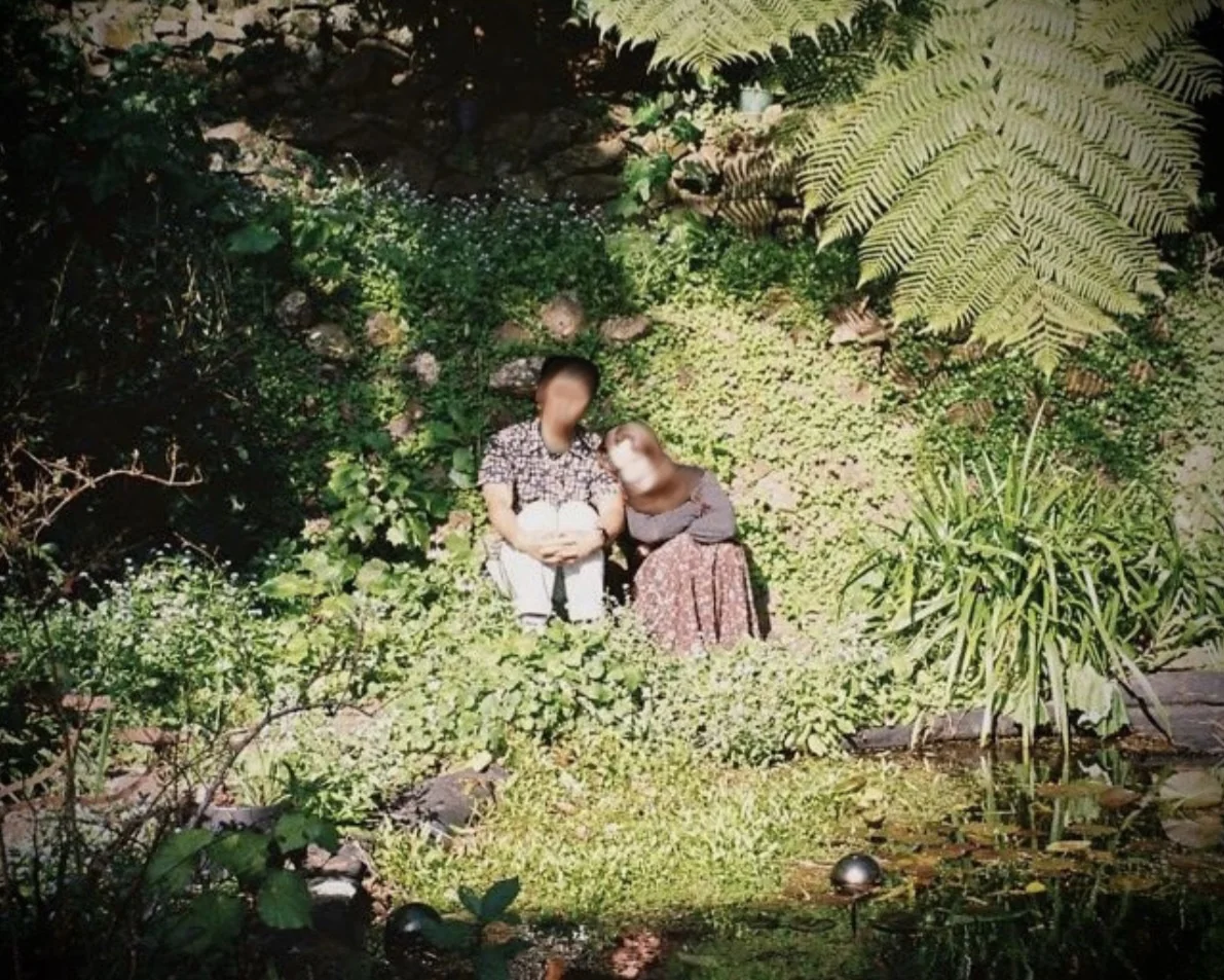 Two figures, faces obscured, seated close together in a sunlit, overgrown garden beside a pond.