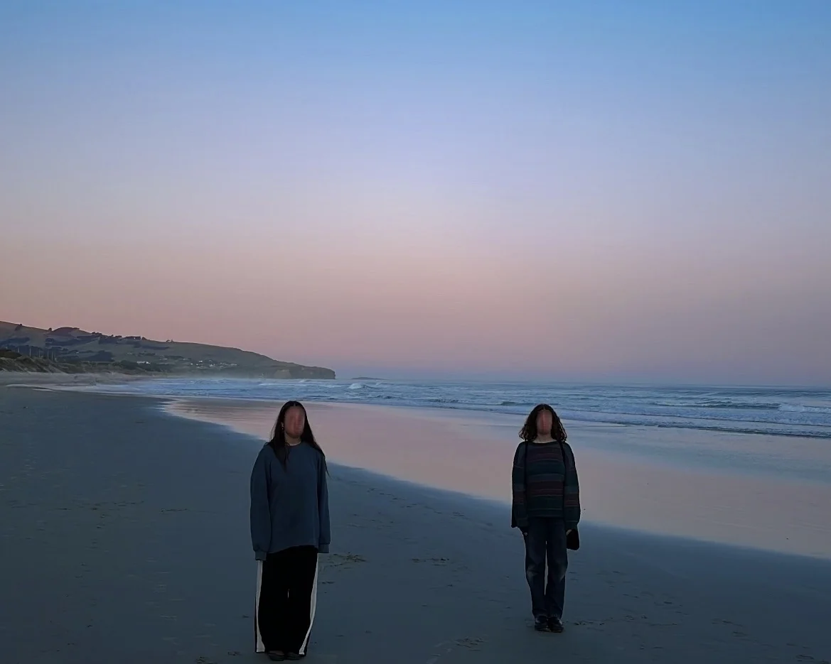 Two figures, faces obscured, standing on an empty beach at dusk beneath a pale blue and pink sky.