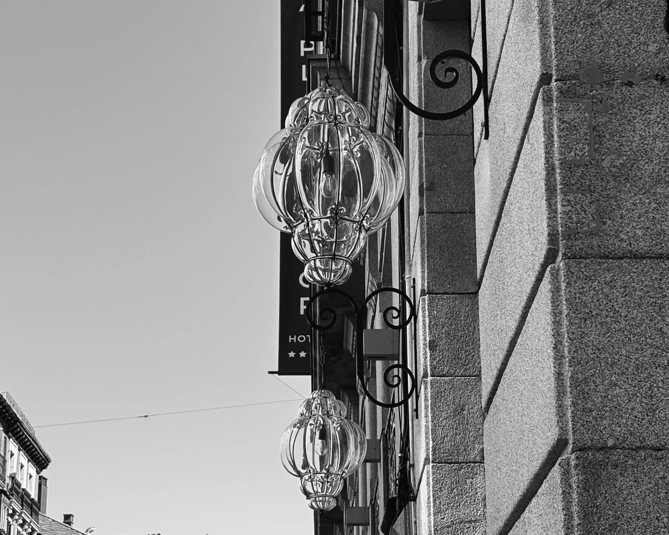Two hanging glass lantern globes on ornate iron brackets against a stone facade, black and white, Madrid