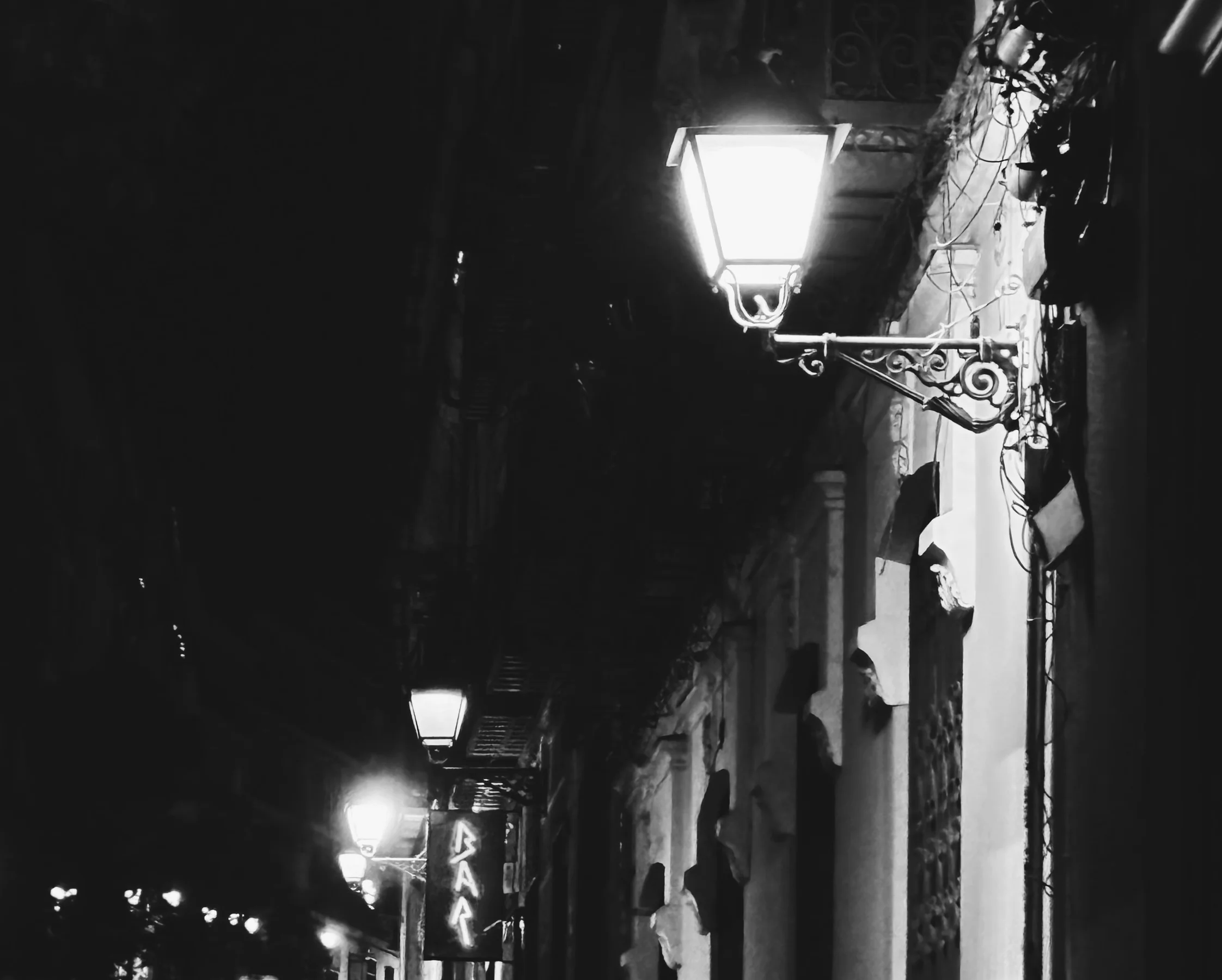 A row of lamps down a dark street, a neon bar sign glowing, black and white, Madrid